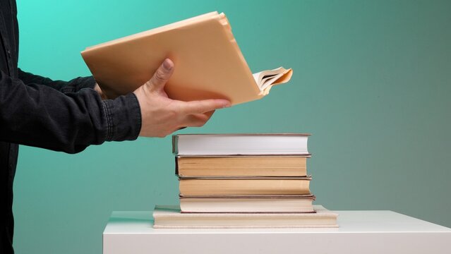 Person in a black shirt holding a manila file folder with papers over a stack of old and new books, symbolizing education, research, and learning on a white surface with a turquoise background