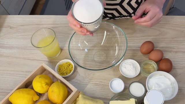 A person pours white granulated sugar from a glass cup into a large clear mixing bowl on a light wooden table surrounded by lemons, eggs, and flour