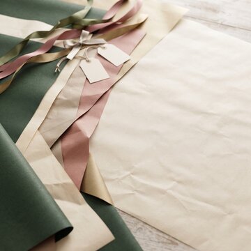 A collection of elegant gift wrapping materials, including textured paper, satin ribbons in pastel hues, and blank gift tags, are artfully arranged on a rustic wooden surface.