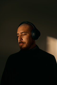 Serene bald man listening to music with closed eyes on headphones in warm sunlight studio portrait, relaxed peaceful expression wearing turtleneck sweater dark dramatic moody light audio meditation.
