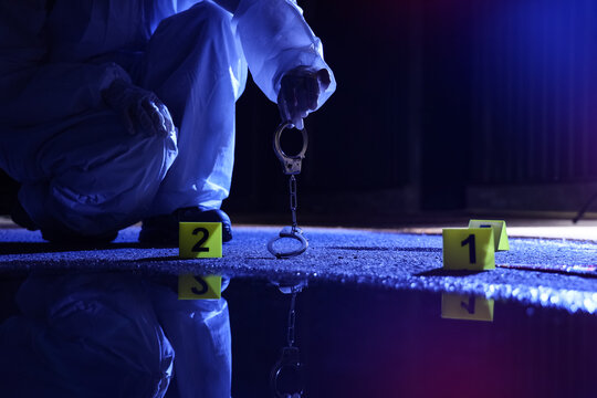 Forensic investigation and crime scene analysis, investigator in protective suit collecting evidence with handcuffs and markers on dark wet ground