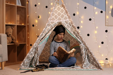 Childhood imagination and creative play, boy wearing pirate hat reading old book inside a cozy indoor teepee tent with fairy lights in a bedroom © Pixel-Shot