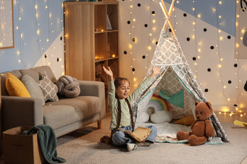 Childhood imagination and storytelling, happy girl reading a book with excitement in a cozy playroom with a teepee tent and warm fairy lights © Pixel-Shot