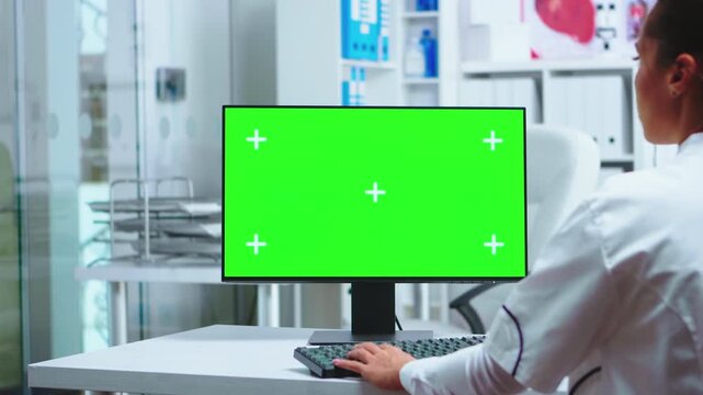 Doctor works at a green screen computer in the clinic while an assistant in uniform stays nearby for healthcare workflow with monitor mockup space.