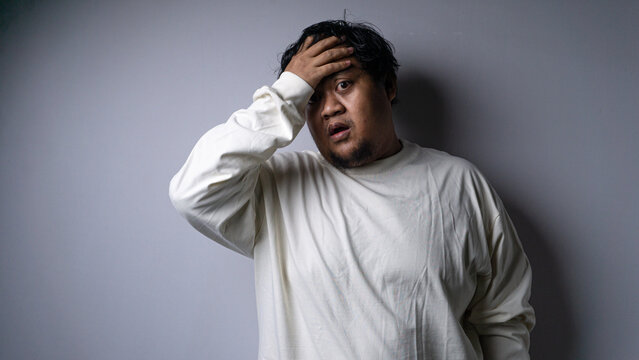 An adult Asian male slaps his forehead with one hand while displaying a shocked and dismayed facial expression. Dressed in a casual cream long sleeve shirt, the subject stands against