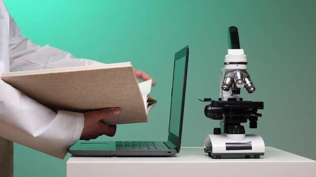 Scientist in lab coat holding an open book, looking up information while working at a laptop next to a laboratory microscope, combining traditional and modern research methods