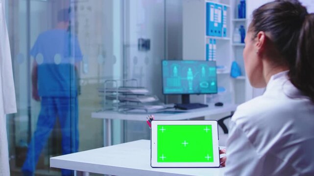 Tablet green screen mockup sits in the foreground of a medical office while a doctor remains at the desk and a colleague in blue scrubs walks away for clinical workflow and screen replacement.