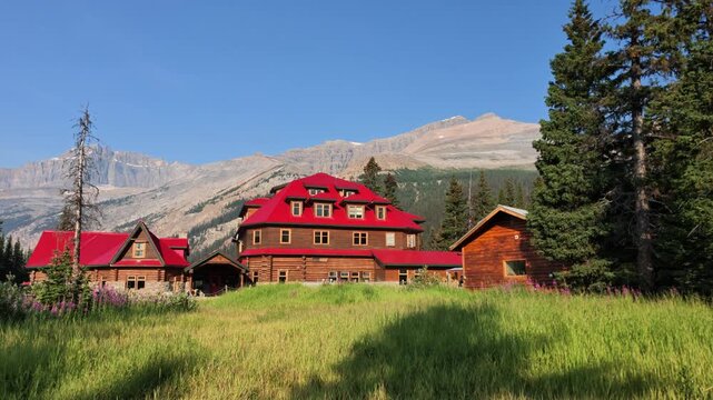 Num-Ti-Jah Lodge and adjacent cabins with vibrant red roofs sit in green meadow, surrounded by pine trees and the towering Rocky Mountains under clear blue sky in Banff National Park, Alberta, Canada.