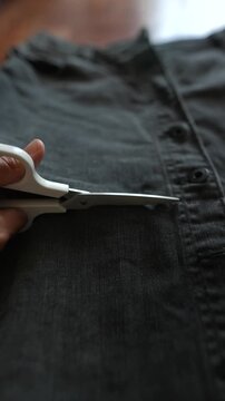 Low close angle view of hand holding white scissors aligning with stitched seam and button row on dark denim jeans in soft indoor light for trimming precisely