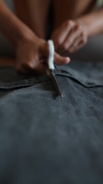Low angle closeup of hands cutting denim seam with white handled scissors on flat work surface, showing blade separation along dark blue stitched fabric texture