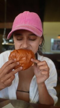 Young woman in pink baseball cap tasting burger with thoughtful side glance, indoor dining close-up, warm soft light, shallow depth of field