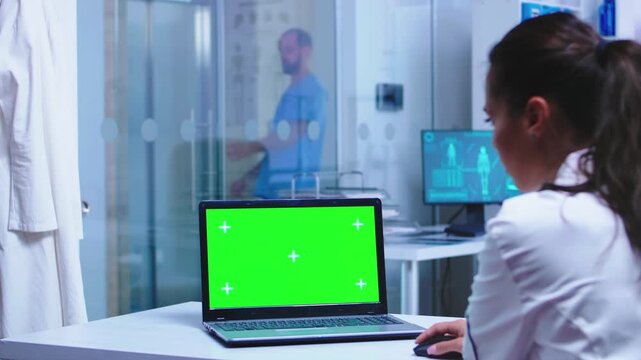 Medical worker uses a laptop with chroma key display while a colleague opens the glass clinic door, creating a practical healthcare technology and clinical workflow scene for screen replacement use.