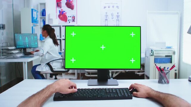 From a first-person perspective, a medical practitioner uses a green screen computer while a doctor works at a nearby clinic workstation for clinical workflow.