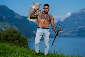 Fototapeta premium Muscular sexy farmer carrying a weathered milk can. Shirtless muscular man with visible chest tattoos. Tattooed muscular torso in a rural portrait. Muscular male model with a traditional pitchfork.