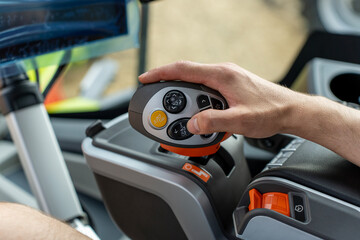 A hand operates a joystick in a heavy equipment cab, with buttons, switches, an armrest, console, and monitor visible. © Marina