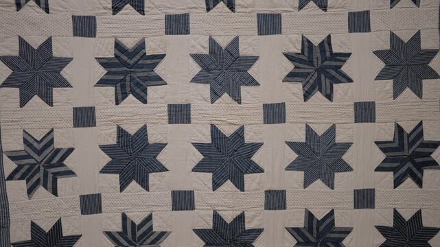 This video shows a panning shot of an old handmade quilt. The camera moves across the vintage textile, showcasing a repeating pattern of geometric blue and white eight-pointed stars and squares.