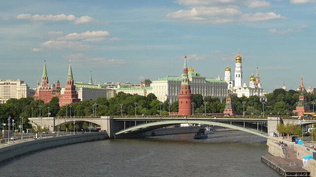 A panoramic view of the historic Moscow Kremlin complex with its towers and palaces, situated along the embankment of the Moskva River under a blue sky with clouds