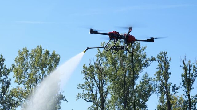 Large multirotor drone spraying a jet of water mist from a long boom nozzle, tethered by a hose, hovering above green trees against a clear blue sky, firefighting and agriculture concept