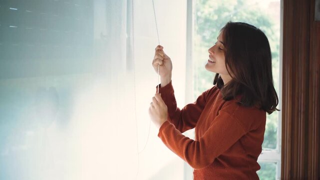 Young Latina woman raising a blackout roller blind at home to let natural light into the room. Concept of home comfort, interior design, natural lighting, privacy, daily routines, and modern home life