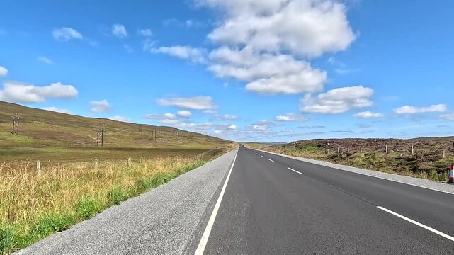 Straight, empty road stretches through rolling, hilly countryside under a blue sky with clouds Heath, mainland, Shetland Islands, Scotland, United Kingdom