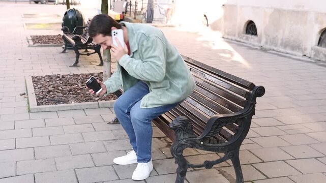 Handsome man with cup of coffee talking on smartphone on bench in city