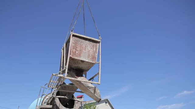 Crane lifts a full concrete bucket above a mixer truck against blue sky, A worker watches the suspended hopper clear the delivery chute on site