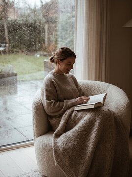 Woman reading a book by the window in a cozy chair at home, wrapped in a soft blanket. Solo Living and Micro Moments express quiet routine, comfort, mindfulness, and peaceful time alone.  