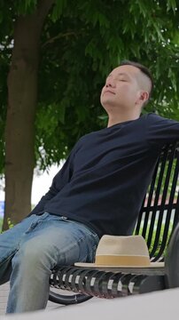 Vertical: Man sits and rests on park bench. He sits on park bench and rests. He takes deep breath under the green trees in the park. Man tries to relax himself by taking deep breaths.