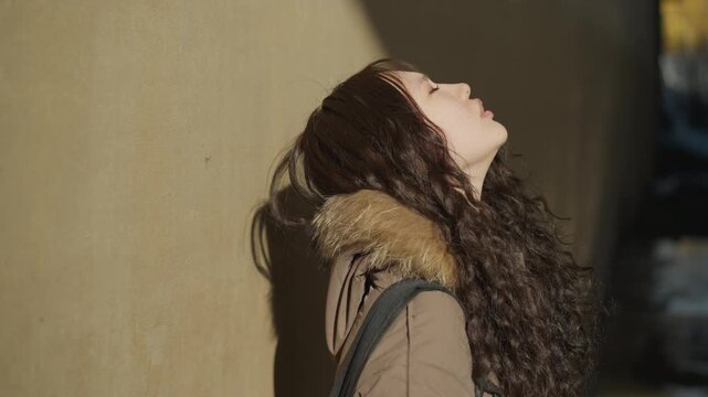 Woman in winter jacket leans casually on bare concrete or plastered urban structure. Her long curly hair and fur trimmed coat suggest cold weather while plain wall behind her provides