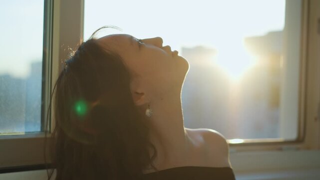 Close up view shows back and partial front of woman with wavy long hair. Woman is positioned near window looking out at urban cityscape during sunset. Soft golden light from setting sun