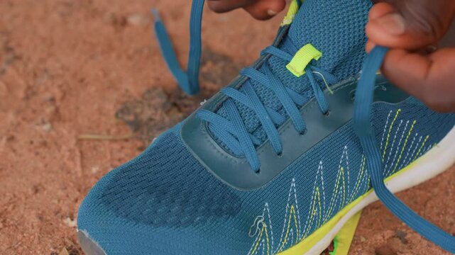 Close up shots show person hand carefully threading lace through eyelets of bright blue sports shoes with yellow accents on rough ground. sequence captures detailed movements as laces are