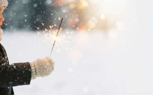 Young child holding a glowing sparkler in winter snow with festive bokeh lights for holiday celebration