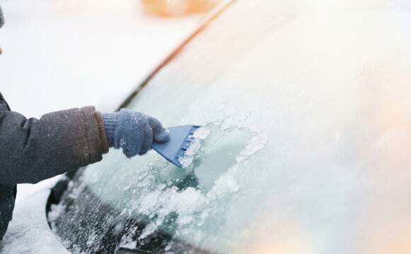Unrecognizable person scraping ice and snow from a car windshield on a cold winter morning representing vehicle maintenance and transportation safety