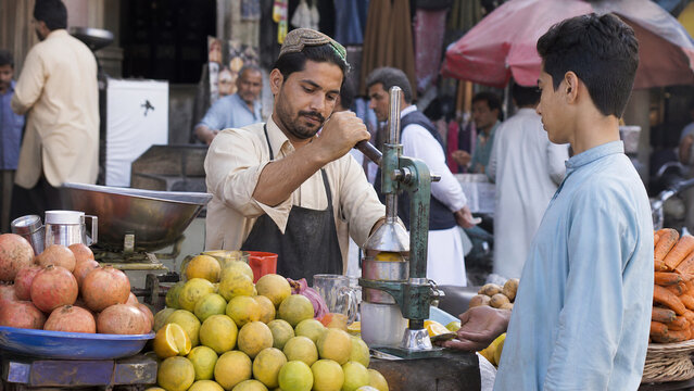 Street food vendor squeezing fresh orange juice at outdoor market stall, male vendor preparing fruit drink for customer