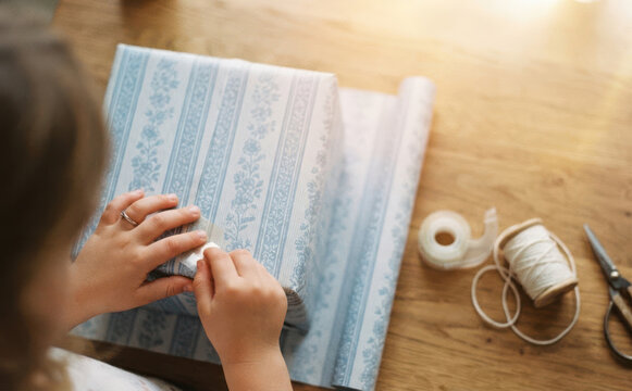 Young girl wrapping a gift box with blue patterned paper at wooden table with tape and string