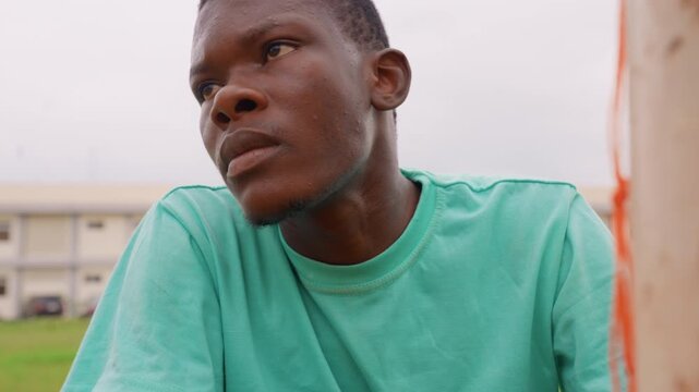 Close up portrait shows young adult male in light green t shirt outdoors. Man is positioned near orange pole with residential buildings and grass visible behind him. shot focuses on his