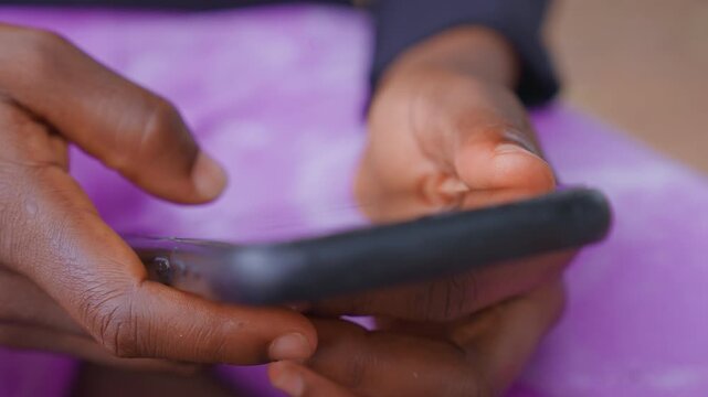 Close up shows two dark skinned female hands holding electronic tablet or phone. background is blurred purple fabric emphasizing texture of skin lines in detail as thumbs press on screen