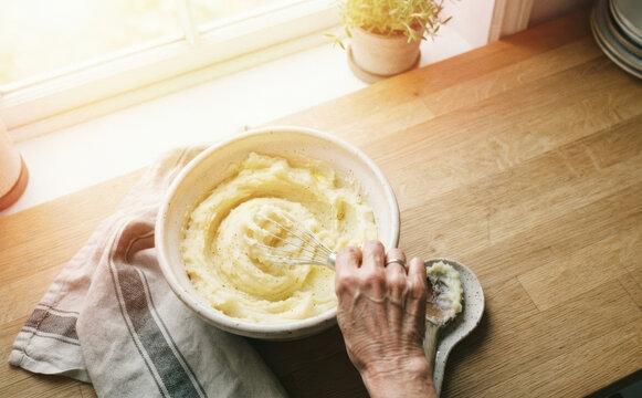 Elderly woman preparing mashed potatoes in ceramic bowl on wooden kitchen counter with whisk and spoon concept of homemade cooking and baking