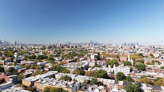 A drone glides over Brooklyn rooftops and tree lined residential streets in New York City. Row homes and large apartment buildings stretch toward the distant skyline beneath the clear blue autumn sky.