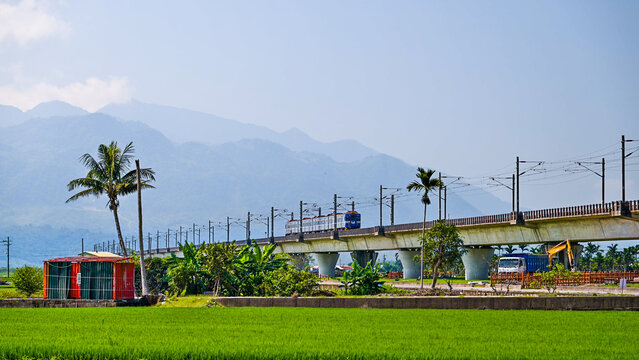 A blue train traveling across a concrete railway bridge in Yuli, Taiwan, with lush green fields, palm trees, and mountain ranges in the background.