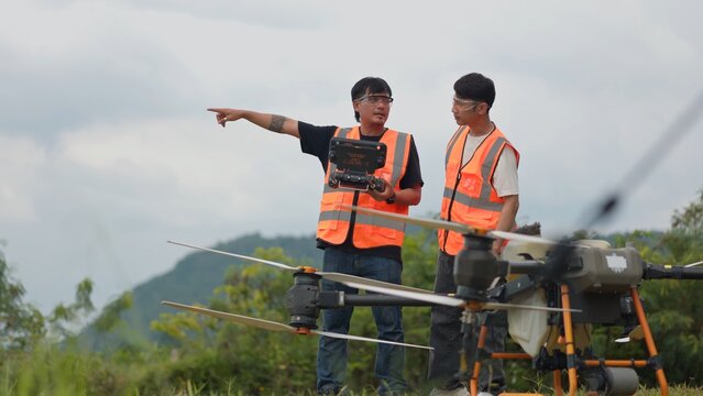 Asian drone pilots in safety vests operating remote controller to fly agricultural spraying drone. Modern smart farming, precision crop fertilization and aerial agritech concept.