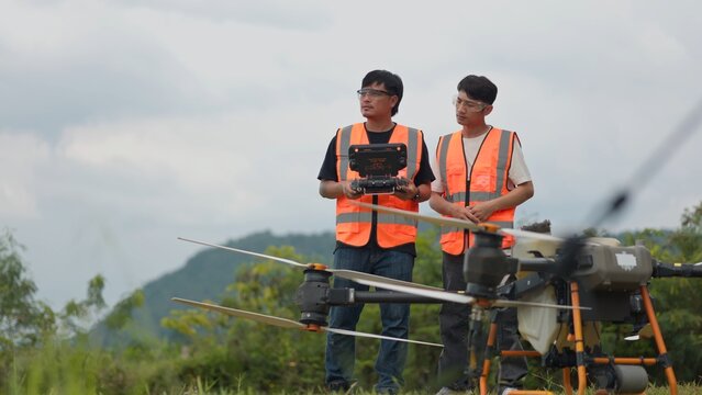 Asian drone pilots in safety vests operating remote controller to fly agricultural spraying drone. Modern smart farming, precision crop fertilization and aerial agritech concept.