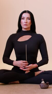 Vertical video. Meditation pose. Zen atmosphere. Woman with black hair sitting in lotus position holding burning incense stick in yoga studio.