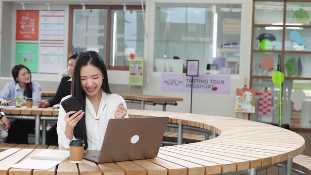 Asian Woman Celebrating Success with Laptop and Smartphone in Bangkok Cafe
