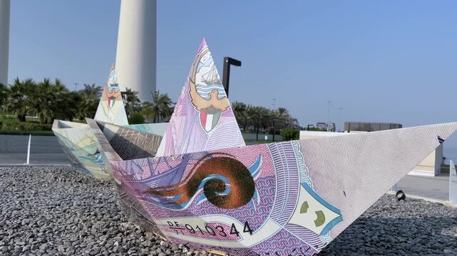 A unique paper boat sculpture decorated with Kuwaiti Dinar banknote graphics sits on a gravel display in Kuwait City during bright daylight. The iconic Kuwait Towers stand in the background.