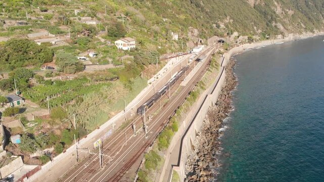 Aerial drone shot of a Trenitalia regional train arriving at Corniglia station, set on the mountain slopes with vineyards on the left and the coast and Mediterranean Sea directly to the right of the r