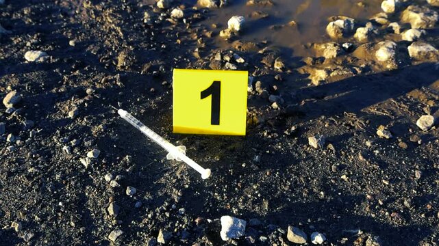 Forensic evidence marker with the number one next to a used syringe on the muddy ground of a crime scene, suggesting drug abuse, overdose, or murder on a gravel road during a sunny day