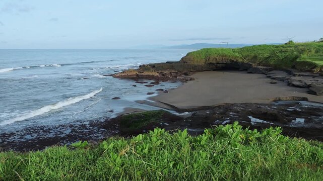 Secluded Bali Black Sand Coastline View With Basalt Rocks And Tidal Pools, Gentle Waves Lapping Empty Beach, Green Headland And Lush Foliage In Foreground, Drone Sweep Capturing Remote Bay