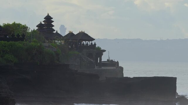Silhouette Tanah Lot Temple Perched On Cliff Overlooking Ocean At Dusk, Multitiered Thatched Roofs, Stone Stairway, Small Figure Standing Near Edge, Misty Horizon, Gentle Waves Breaking Against Rocks,