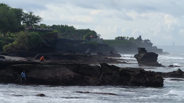 Rugged Rocky Coastline With Lone Fisher, Orangeclad Fisher On Tidal Rock Platform, Waves Crashing Against Basalt Outcrops, Distant Balinese Temple Silhouette Shrouded In Mist, Overcast Sky, Tanah Lot